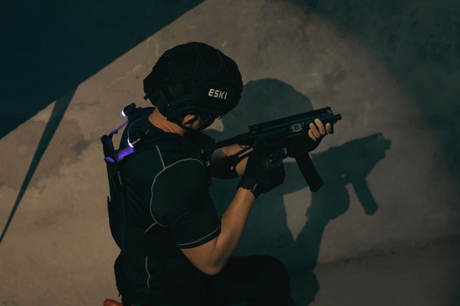 Tactical operator in black Eski gear crouched against concrete wall with submachine gun and helmet in low-light training environment